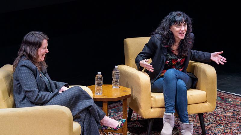 Two people sitting on a dark stage in large comfortable camel-tan chair speaking to each other and the audience.